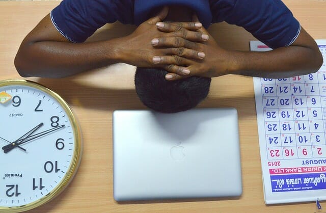 przemęczony mężczyzna trzyma głowę na biurku, obok laptop, kalendarz, zegar